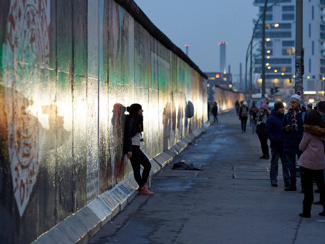 Personen stehen bei Abendlicht an der Mauer der East Side Gallery, eine Person lehnt sich an der Mauer an