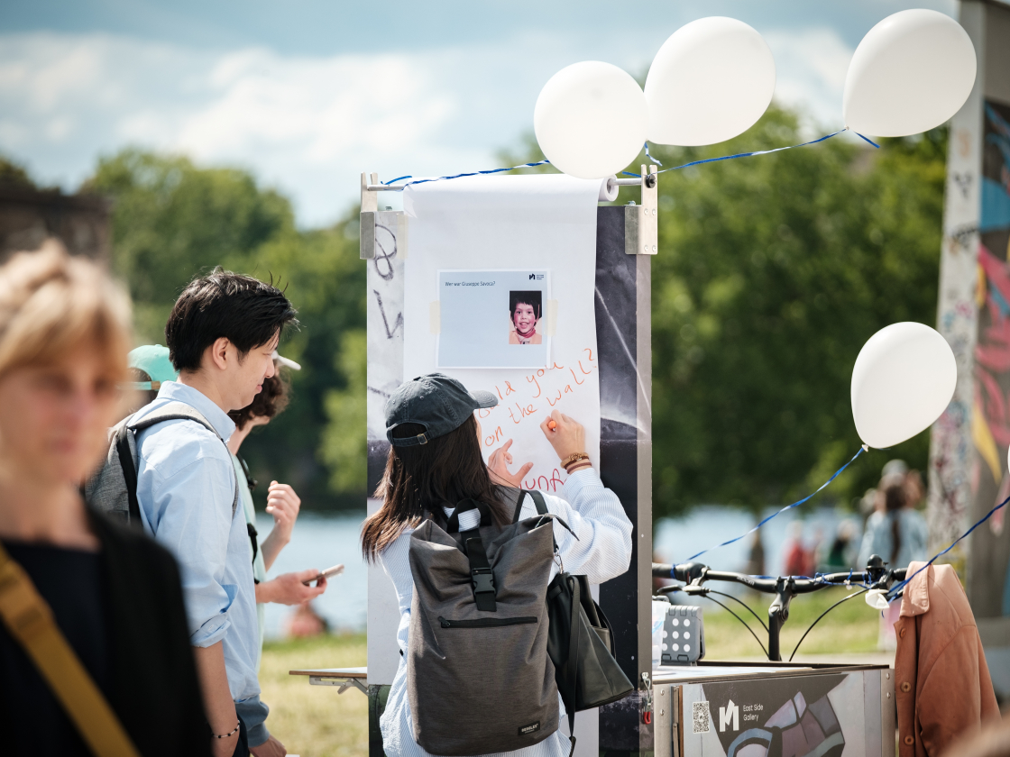 Menschen stehen vor einer Stellwand und schreiben etwas, im Hintergrund Ballons und die East Side Gallery